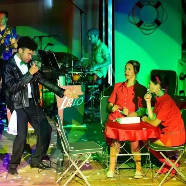 Schauspieler steht auf der Bühne und singt, zwei rot gekleidete Schauspielerinnen schauen zu 