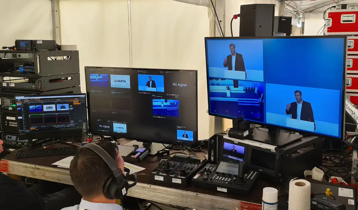 Produktion Eine Person mit Kopfhörern sitzt an einem Arbeitsplatz mit mehreren Monitoren, auf denen ein Mann zu sehen ist, der an einem Rednerpult spricht. Der Raum ist voller audiovisueller Geräte und Kabel, was auf eine Live-Übertragung oder eine Veranstaltungsproduktion hindeutet.