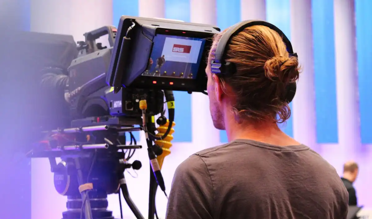 Eine Person mit Kopfhörern bedient in einem Studio eine große Videokamera und filmt eine Szene, die auf dem Kameramonitor zu sehen ist. Im Hintergrund sind blaue und violette vertikale Paneele zu sehen.