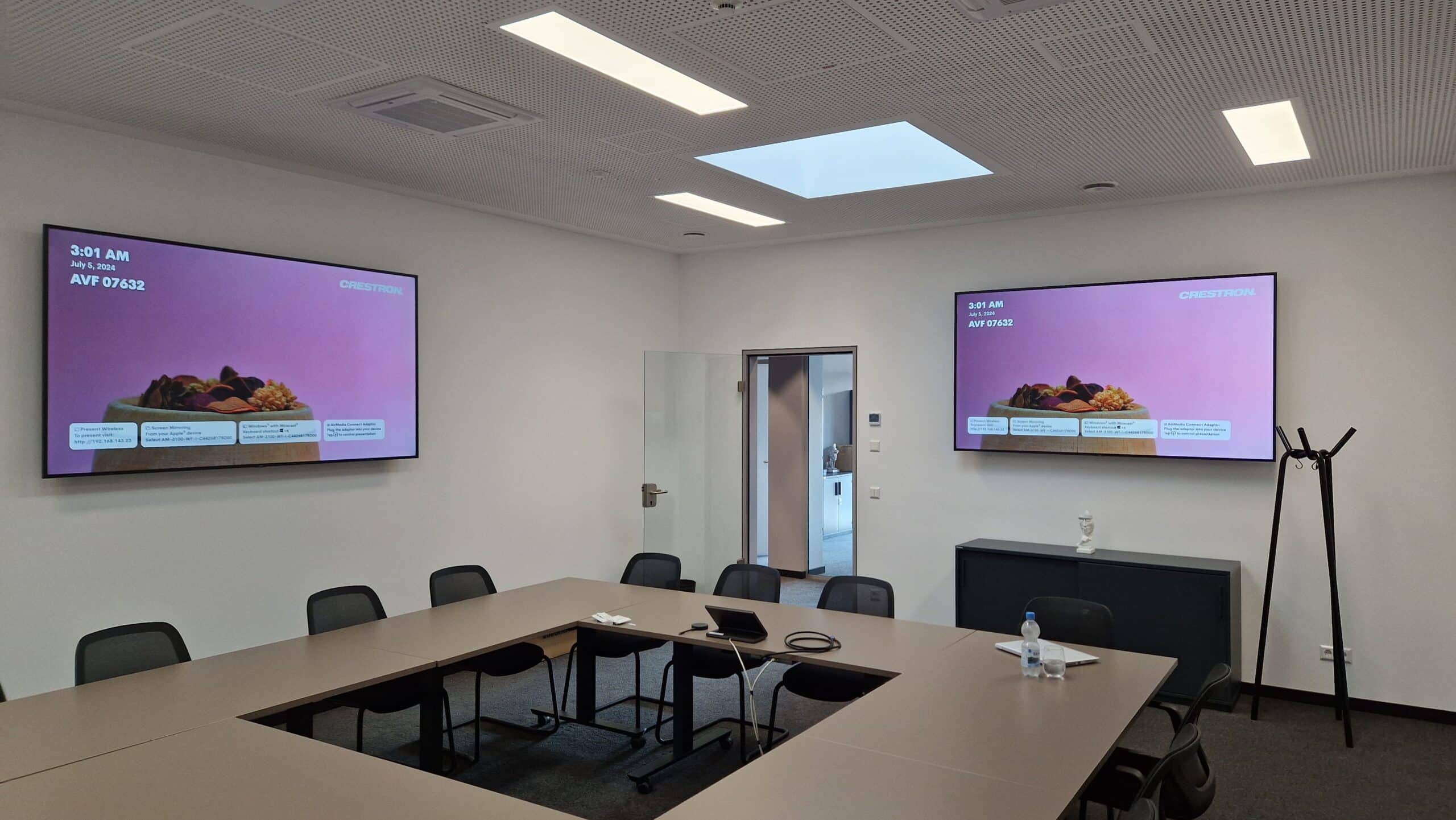 20240813_085451 Ein Konferenzraum mit einem U-förmigen Tisch, schwarzen Stühlen, zwei Bildschirmen an der Wand, auf denen identische Inhalte zu sehen sind, einer Garderobe, einer Wasserflasche auf dem Tisch und einem Laptop in der Mitte des Raums.