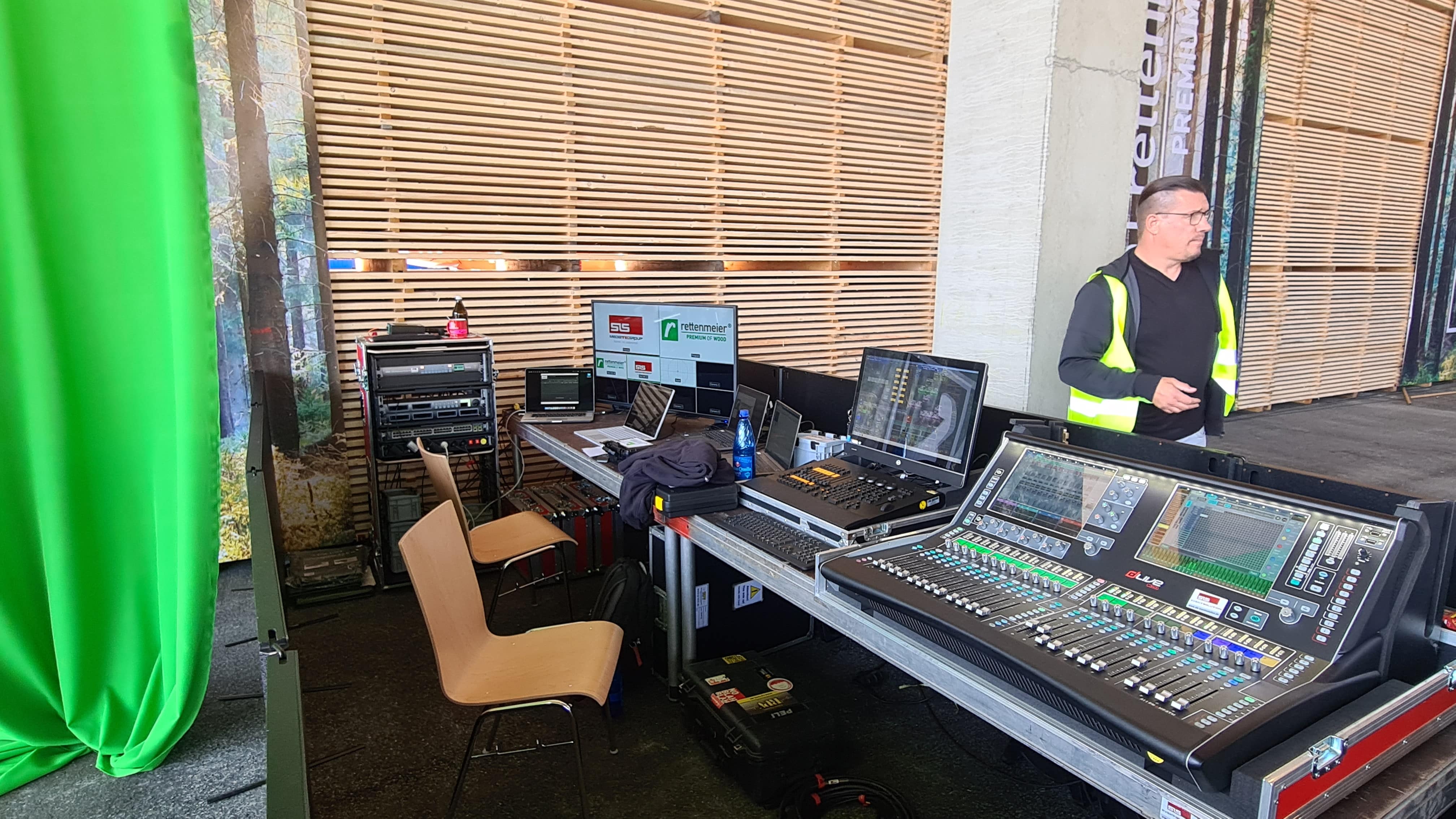 Eine audiovisuelle Steuerungsanlage mit Mischpulten, Monitoren und Geräten in einem Innenraum. Zwei Stühle und ein Greenscreen sind zu sehen. Ein Mann in einer Warnweste steht daneben.