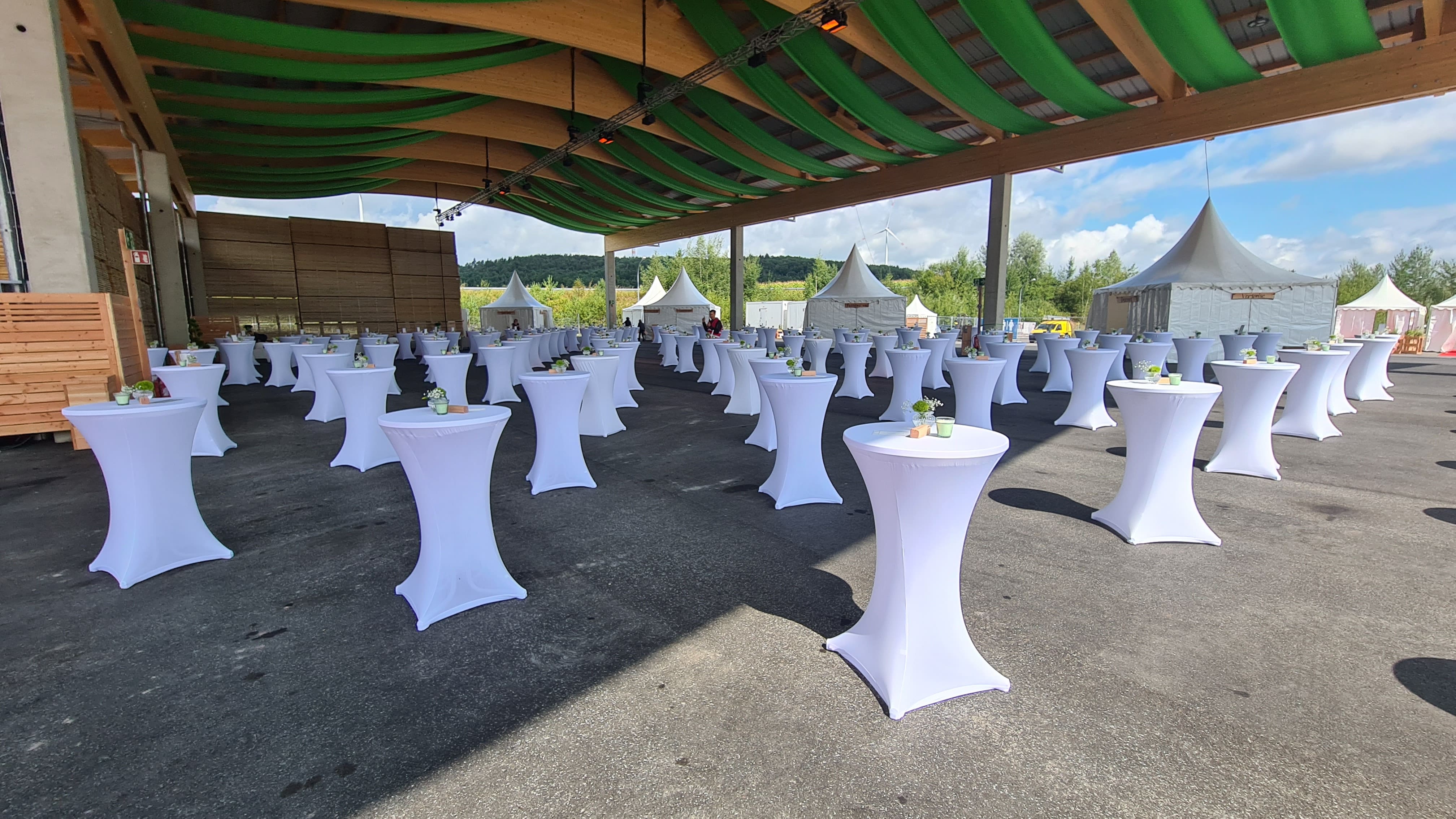 An einem sonnigen Tag stehen Reihen weißer Cocktailtische mit kleinen Blumengestecken unter einem großen überdachten Außenbereich mit grün gestreiftem Deckenstoff, daneben weiße Zelte.