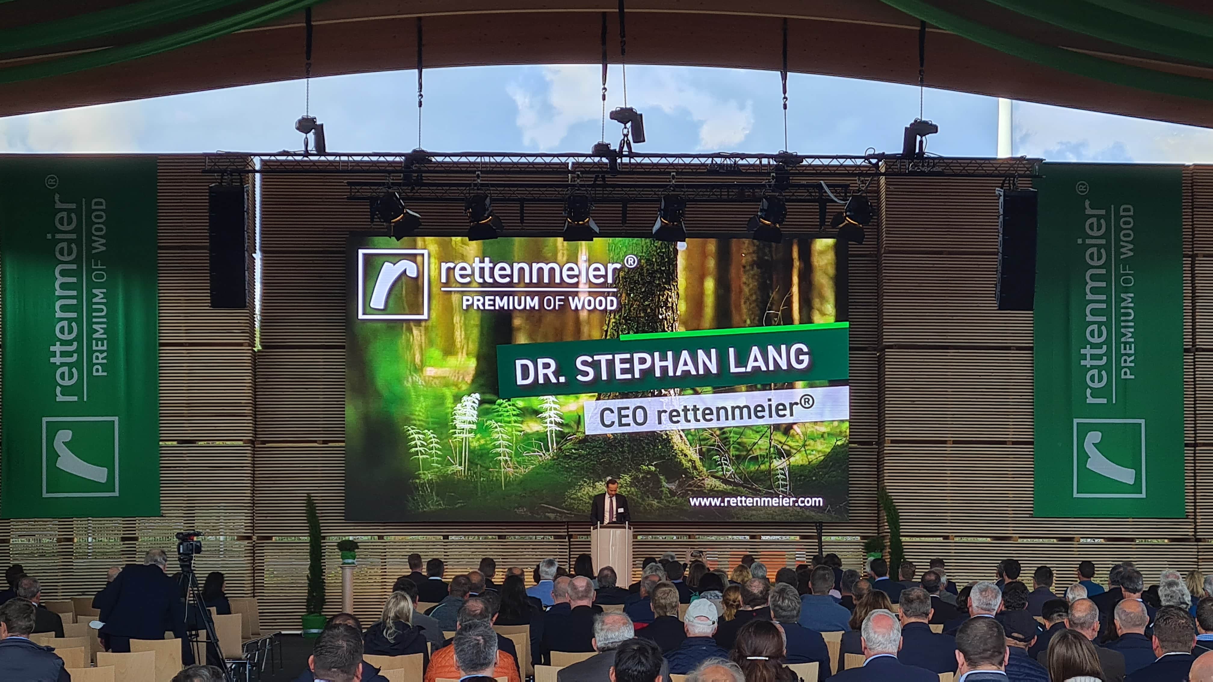 Ein Redner steht auf einem Podium auf einer mobilen Bühne, hinter ihm ist Dr. Stephan Lang, Geschäftsführer von rettenmeier, auf einem großen Bildschirm zu sehen, und spricht vor Publikum bei einer Antenne Bayern SchulhofGIG-Veranstaltung in der Halle.
