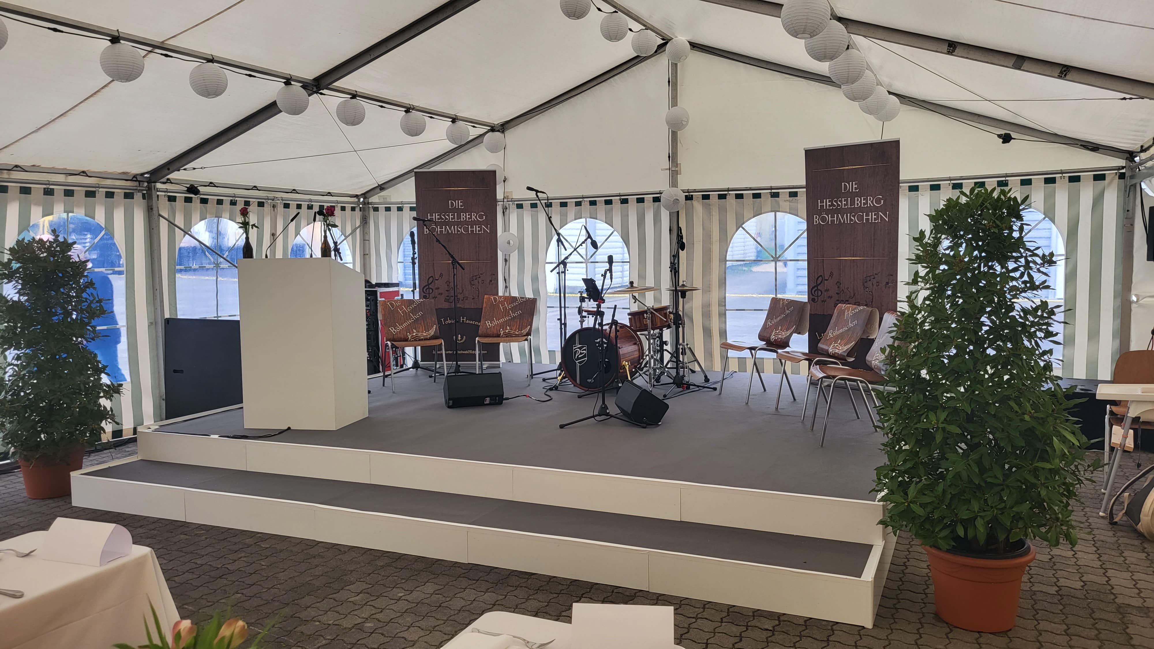 Ein weißes Veranstaltungszelt mit einer Bühne, auf der ein Schlagzeug, Stühle, zwei große braune Banner, Topfpflanzen und weiße Laternen, die von der Decke hängen, zu sehen sind. Vor der Bühne stehen Tische mit weißen Tischdecken.