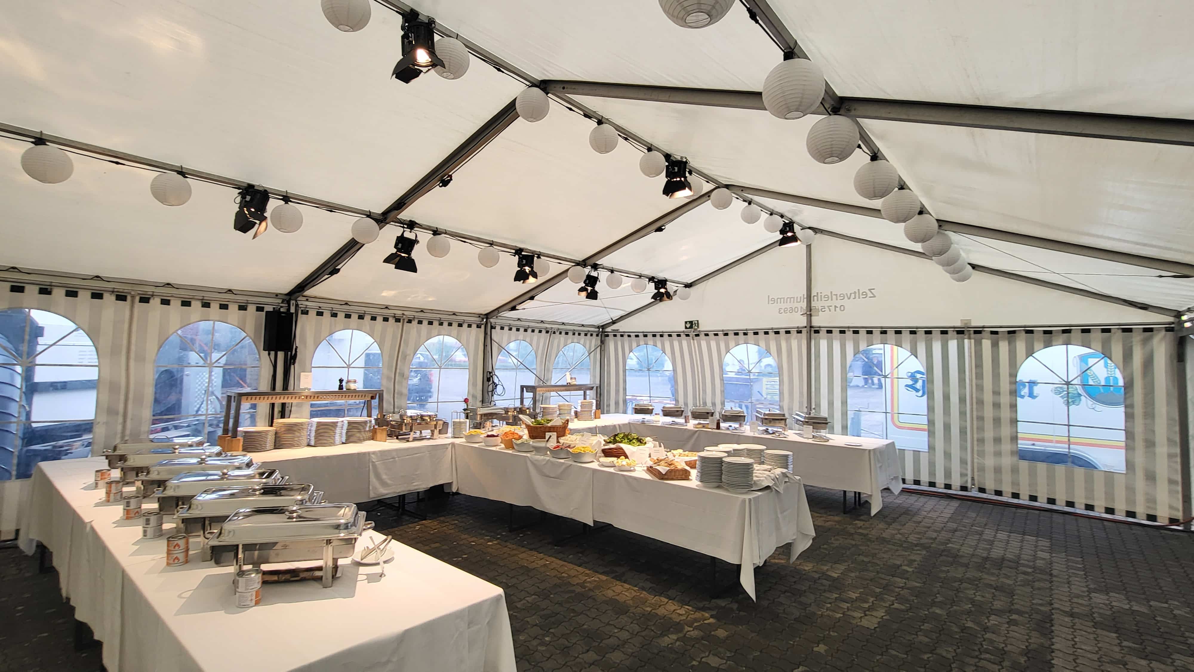 Ein Catering-Bereich in einem großen weißen Festzelt. Mehrere lange, mit weißen Tischdecken gedeckte Tische sind in U-Form angeordnet; darauf stehen Edelstahl-Warmhaltebehälter, Stapel von Tellern und verschiedene Schüsseln mit Speisen. An der Decke hängen runde Papierlaternen.