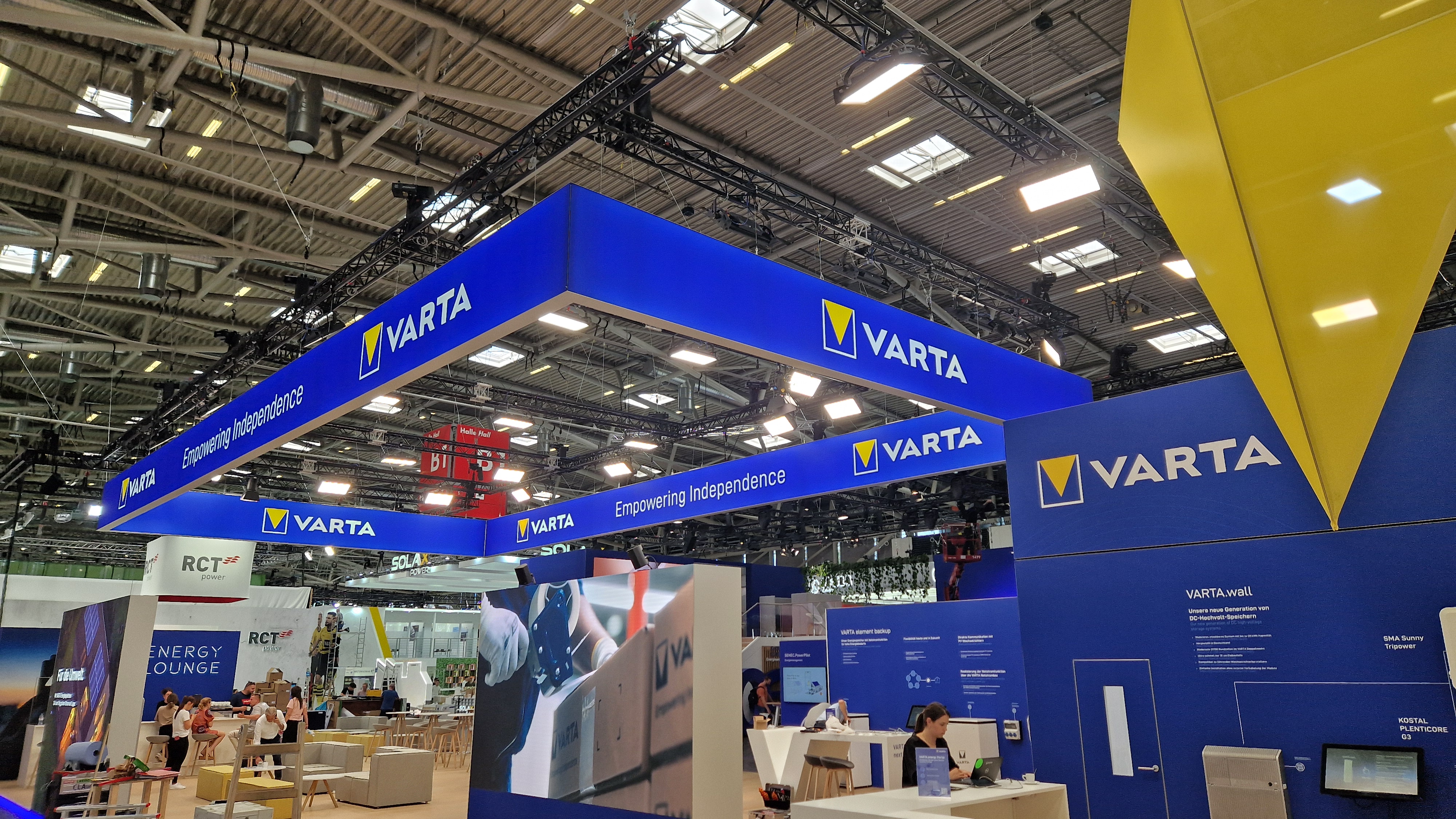 Ausstellungshalle mit einem großen blauen VARTA-Stand, auf dem Markenlogos und der Slogan „Empowering Independence“ zu sehen sind. Unter hohen Deckenleuchten sind Menschen zu sehen, die an verschiedenen Arbeitsstationen beschäftigt sind.