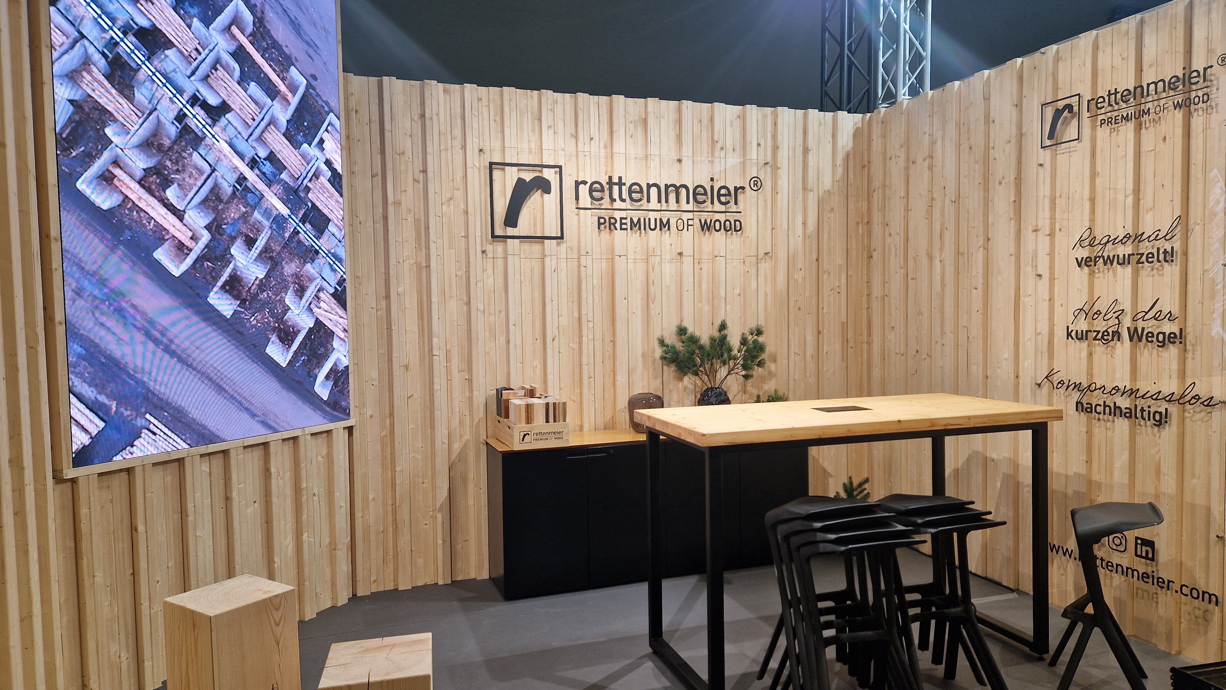 Messestand im Holz-Design mit hellen Holzwänden, einem Stehtisch mit schwarzen Hockern, einer kleinen Pflanze, Aufbewahrungsschränken und einem großen Bildschirm, auf dem ein Bild eines Holzstapels aus der Vogelperspektive zu sehen ist. Das „Rettenmeier Premium of Wood“-Logo ist gut sichtbar.