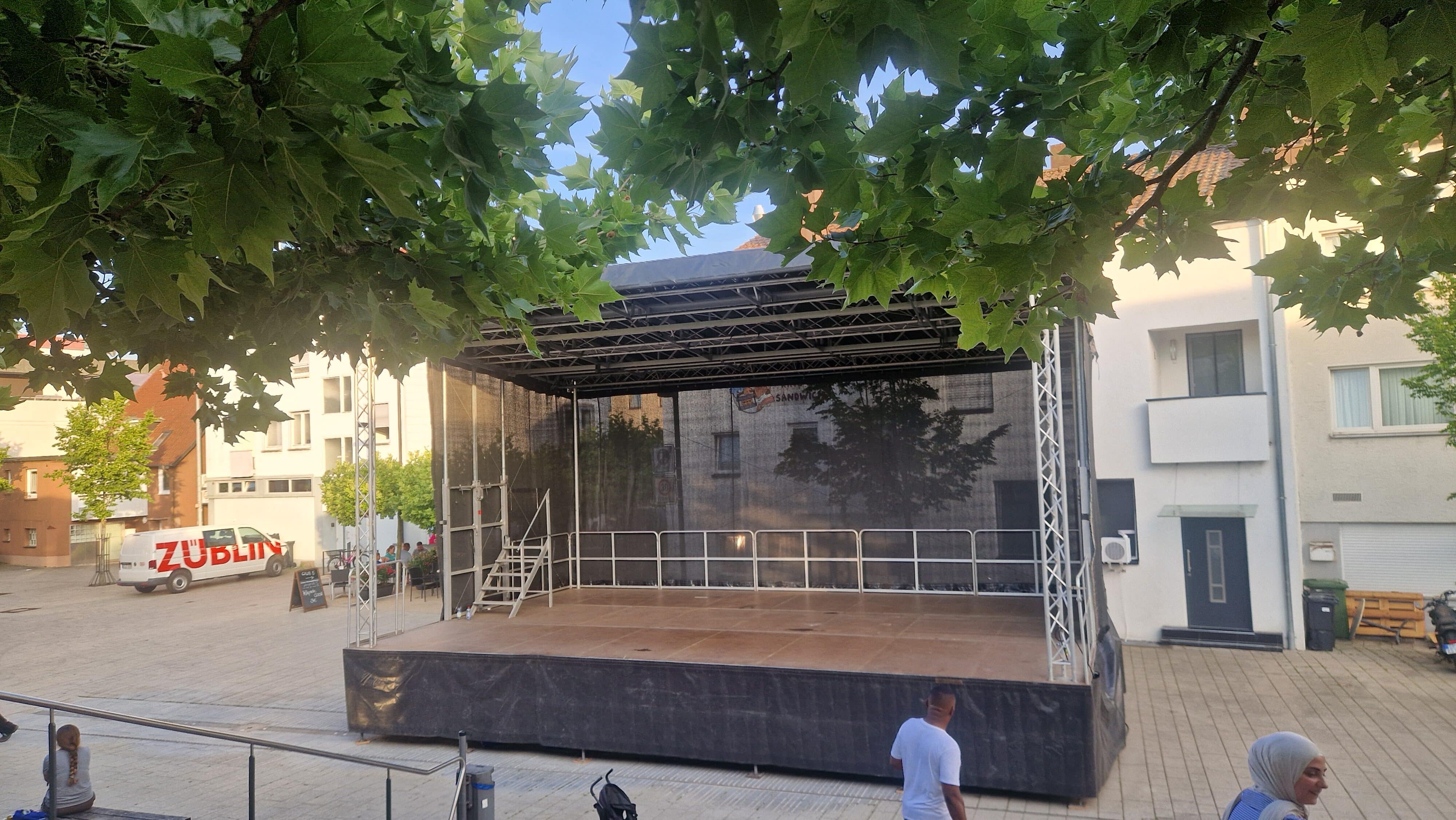 Auf einem Platz steht eine leere Freiluftbühne mit schwarzen Vorhängen, umgeben von Gebäuden und Bäumen. Im Hintergrund sind einige Menschen und ein Lkw zu sehen.