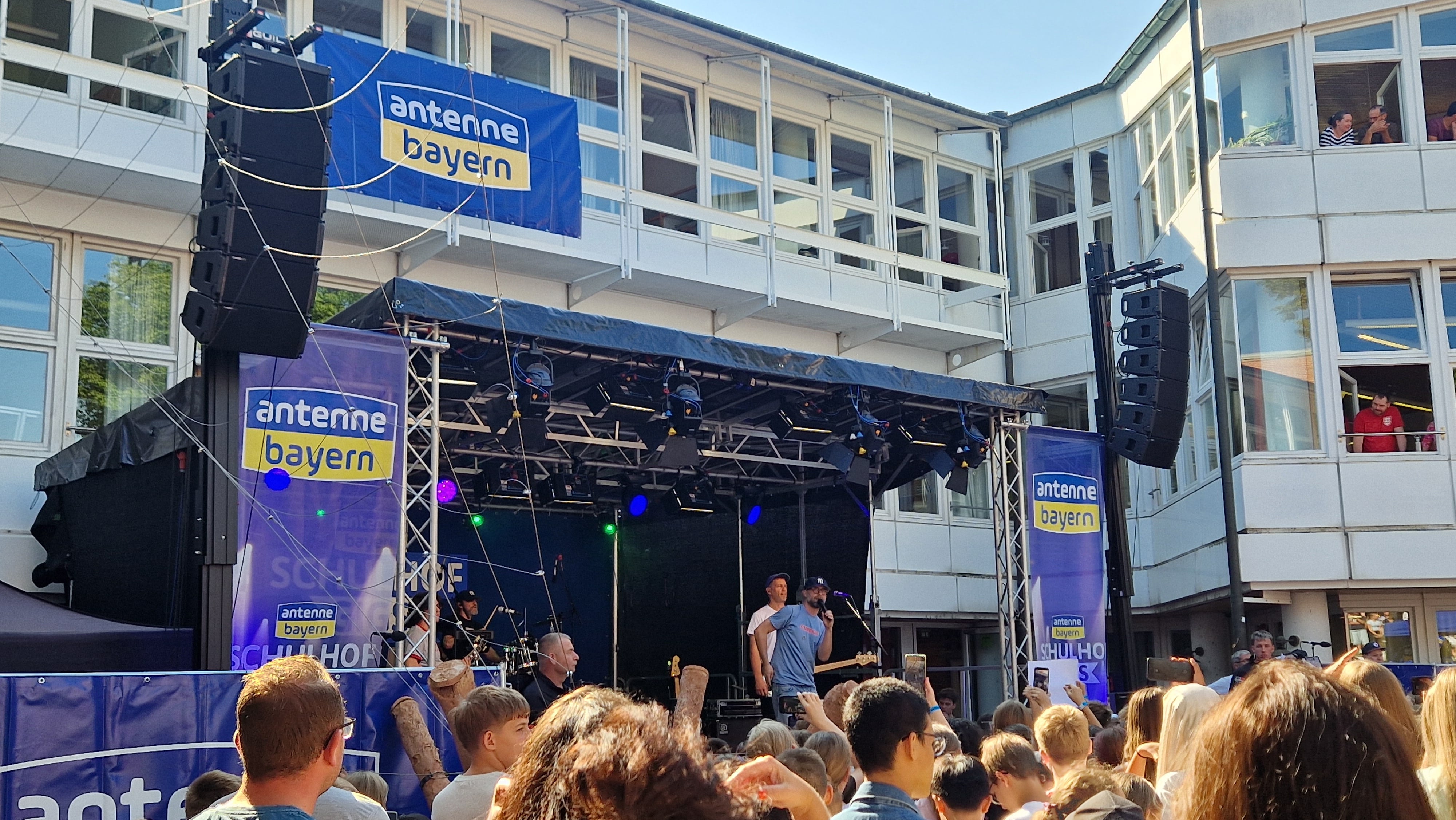 Eine Menschenmenge verfolgt ein Live-Konzert unter freiem Himmel auf einer Bühne mit Antenne-Bayern-Bannern, umgeben von weißen Gebäuden und blauem Himmel.