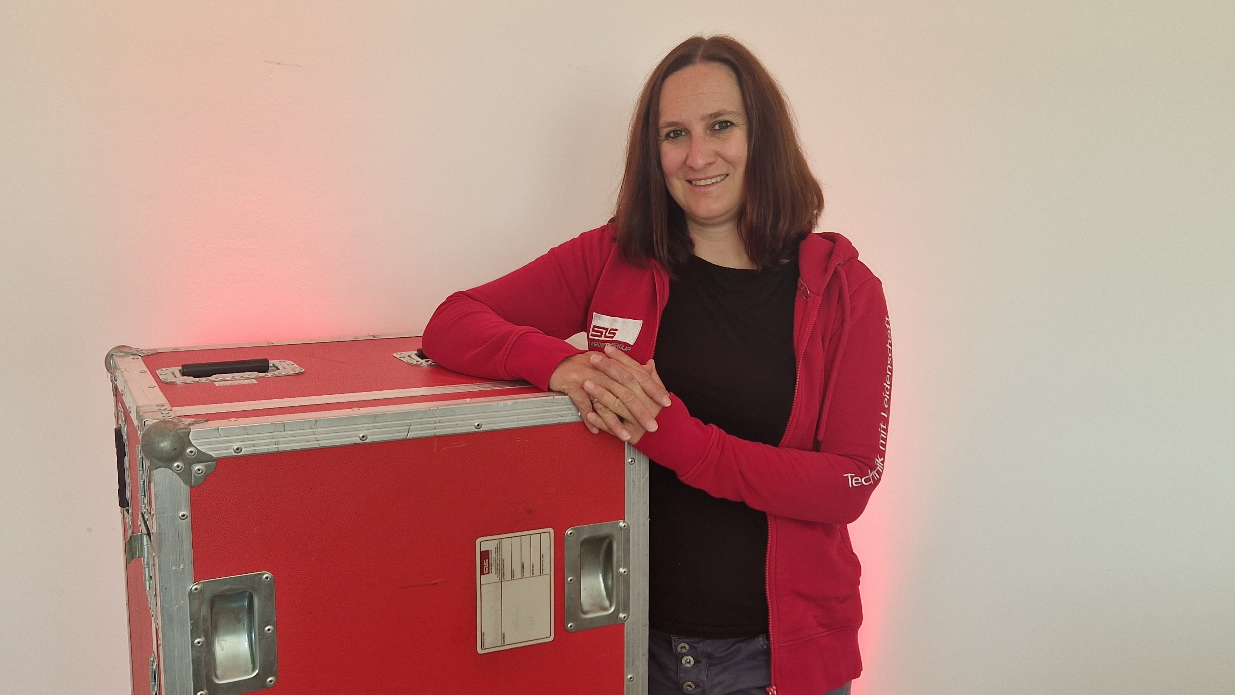 Eine Frau in einem roten Kapuzenpulli lehnt an einem großen roten Instrumentenkoffer, der an einer weißen Wand steht, und lächelt in die Kamera.
