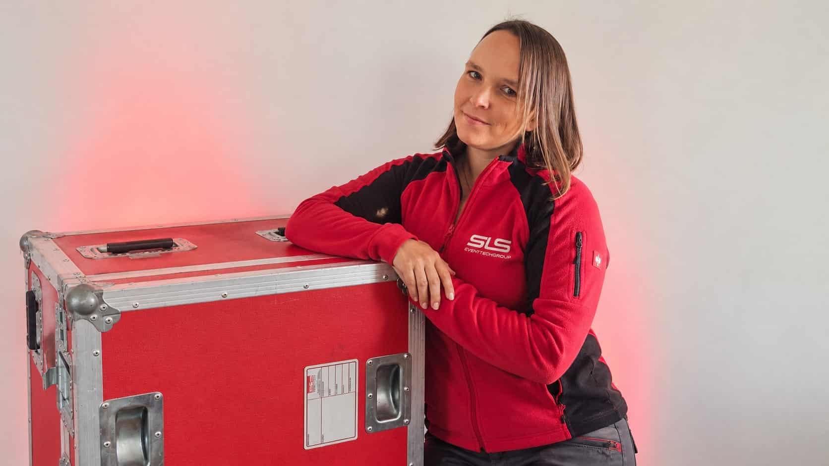 Eine Frau in einer rot-schwarzen Jacke mit der Aufschrift „SLS“ lehnt sich vor einem hellen Hintergrund an einen großen roten Gerätekasten.