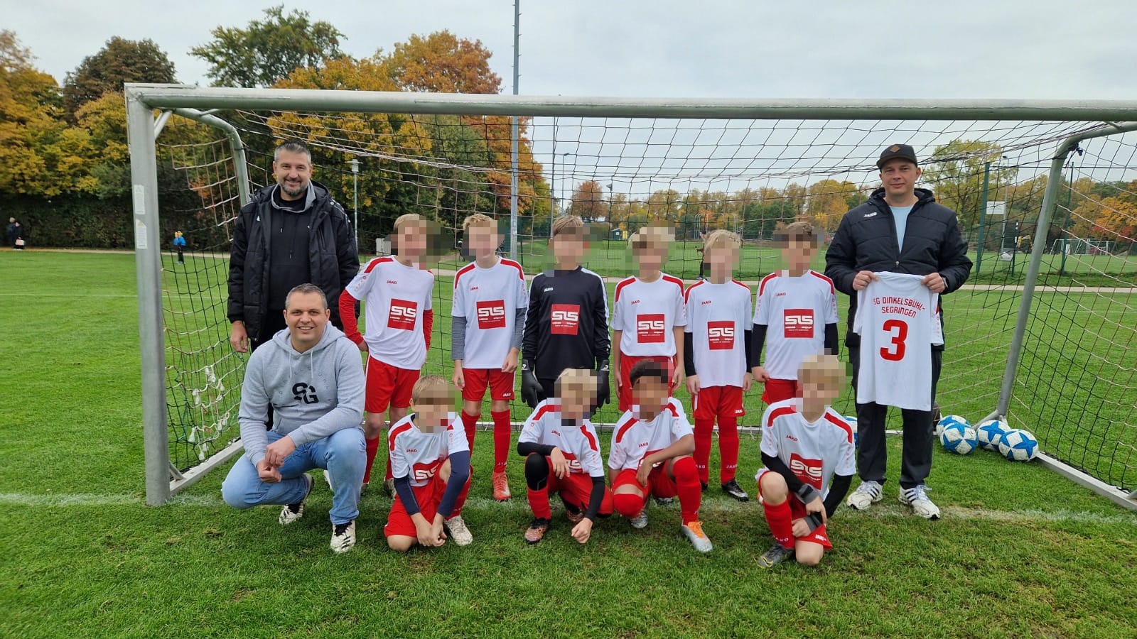 WhatsApp Image 2026-09-24 at 16.28.58 Eine Jugendfußballmannschaft in weißen und roten Trikots posiert zusammen mit drei Erwachsenen vor einem Tor. Ein Erwachsener hält ein Mannschaftstrikot in der Hand. Auf dem Rasen daneben liegen blaue und weiße Fußbälle. Im Hintergrund sind Bäume und eine Wiese zu sehen.